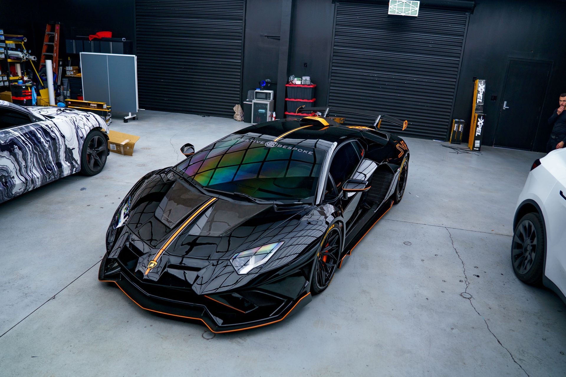 Black sports car in a garage with a colorful reflective windshield, flanked by other cars.