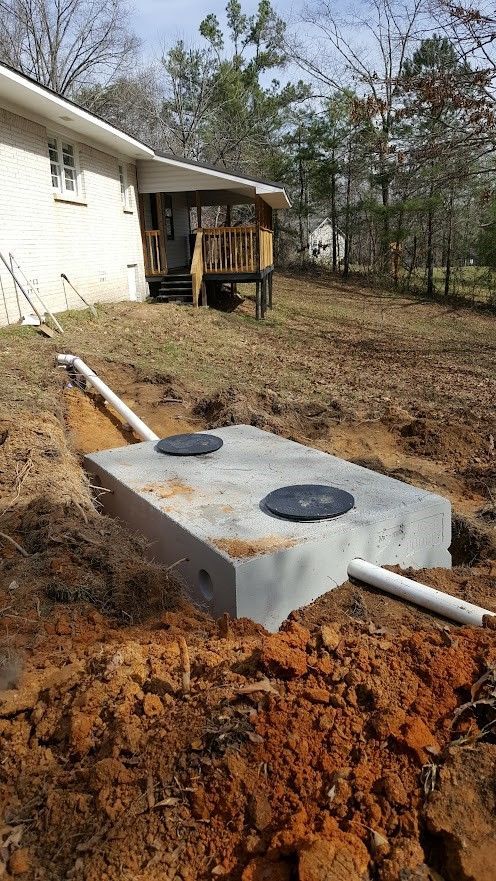 A rectangular concrete septic tank in an excavated dirt pit near a house. Two black covers are visible on top, and white PVC pipes connect to it. A rectangular concrete septic tank in an excavated dirt pit near a house. Two black covers are visible on top, and white PVC pipes connect to it.
