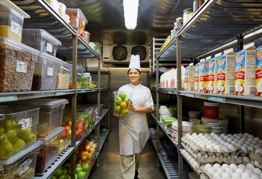 Person in a chef uniform standing in a stocked walk-in pantry, holding vegetables and smiling.
