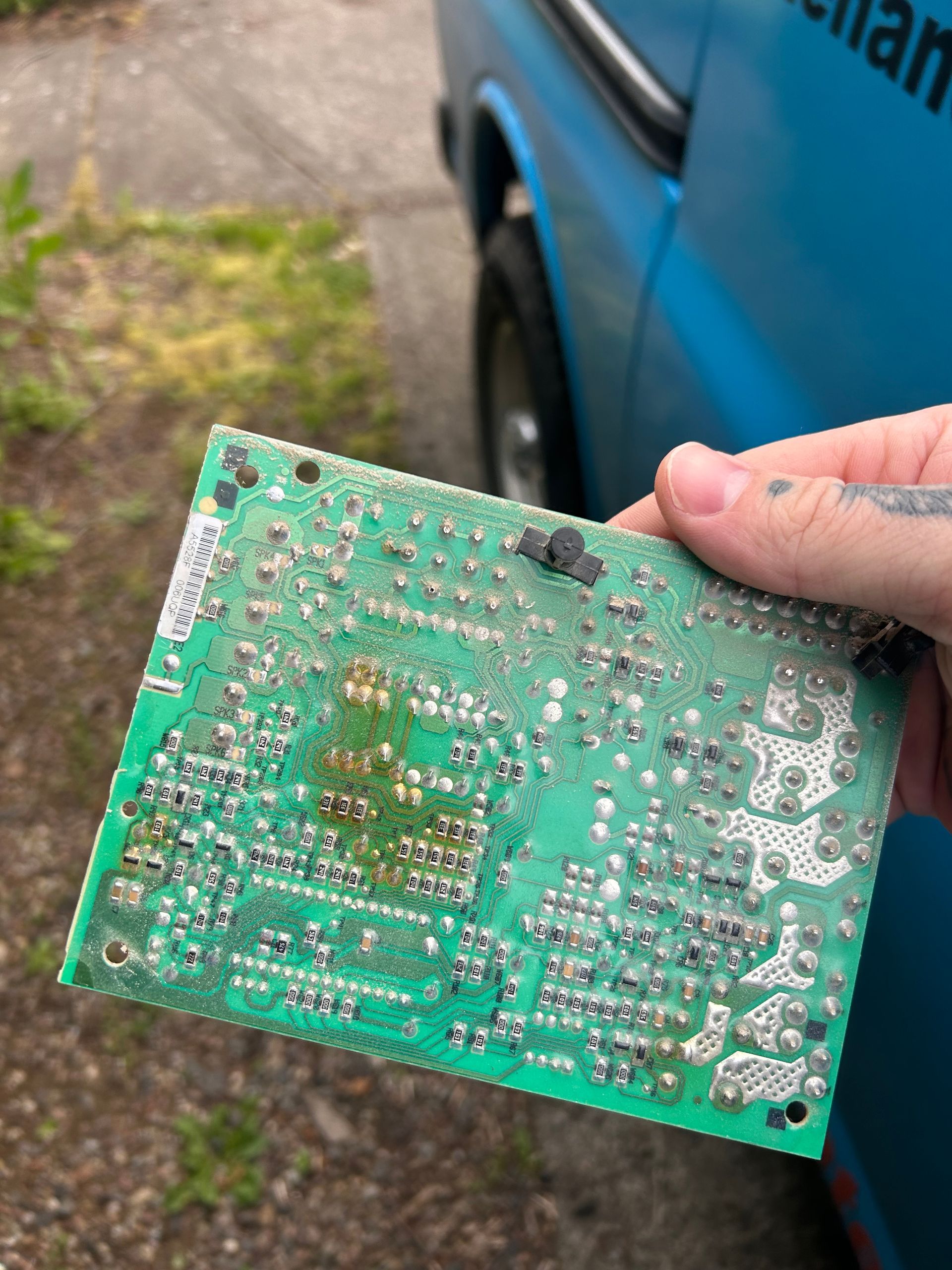 Hand holding a green circuit board in front of a blue vehicle outdoors