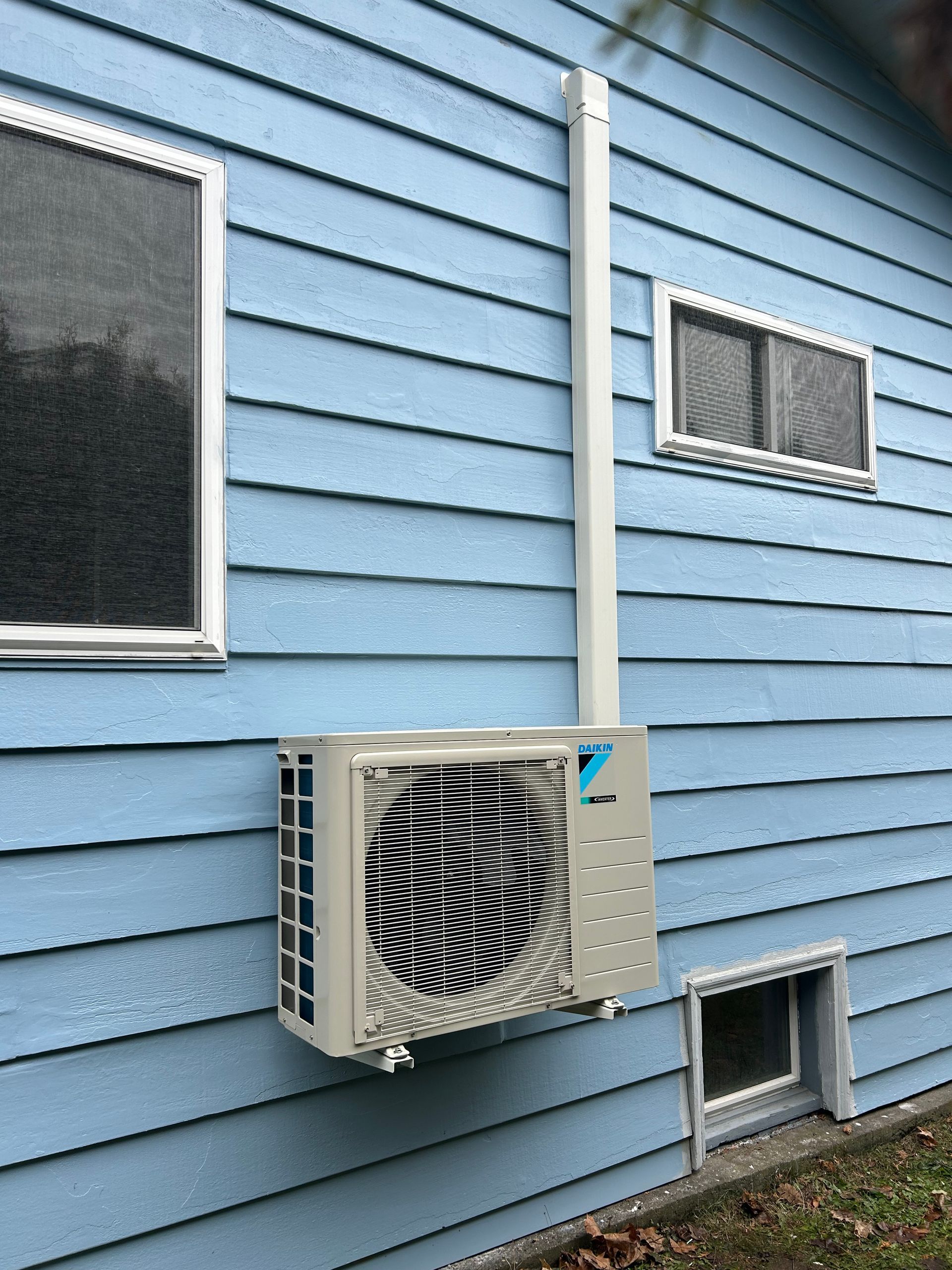 Outdoor air conditioner unit mounted on a light blue house wall beneath two windows.