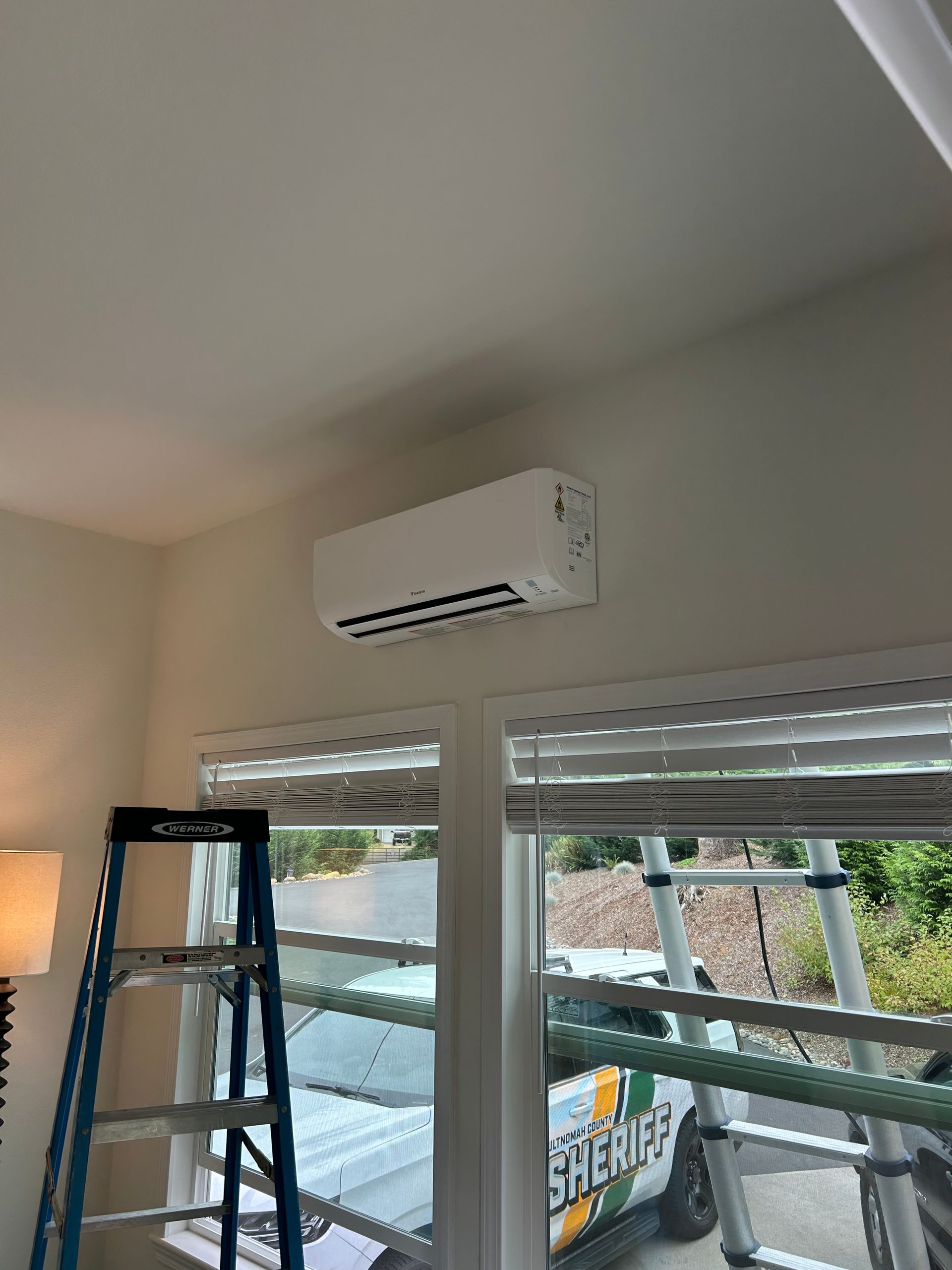 Wall-mounted air conditioner above windows in a room, with a ladder and floor lamp nearby.