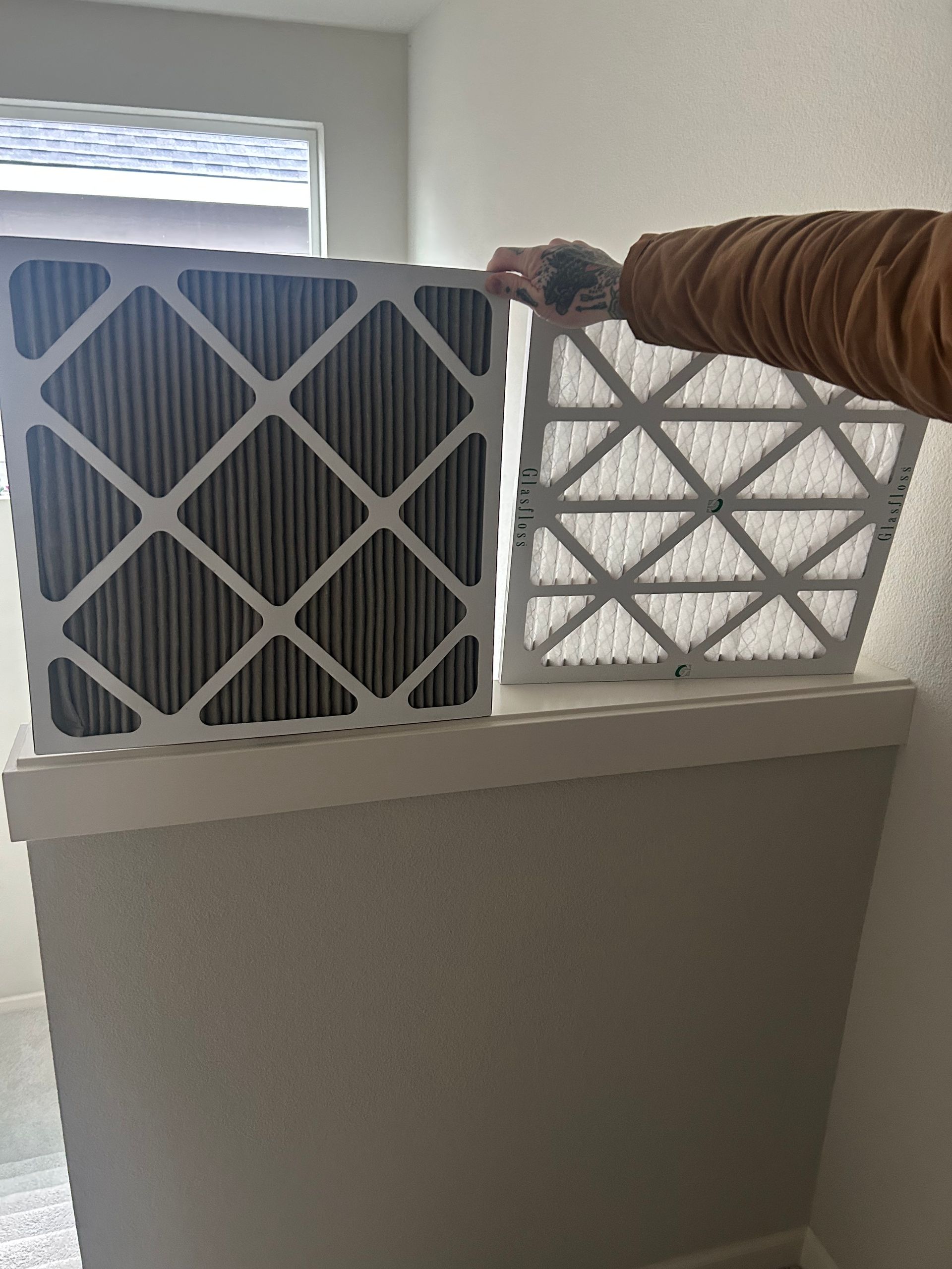 Person holding a white air filter above a vent, with another filter beside it on a ledge
