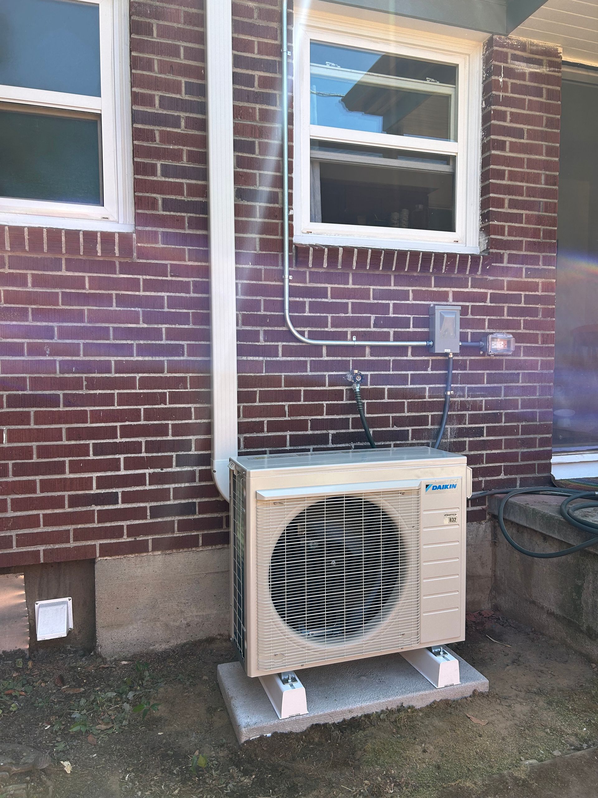 Outdoor air conditioner unit beside a brick house, with white downspout and windows above.