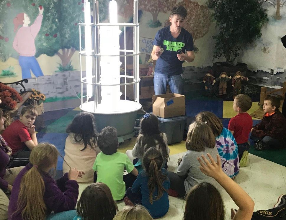 Children seated on the floor watch a man speaking beside a lit display in a classroom.