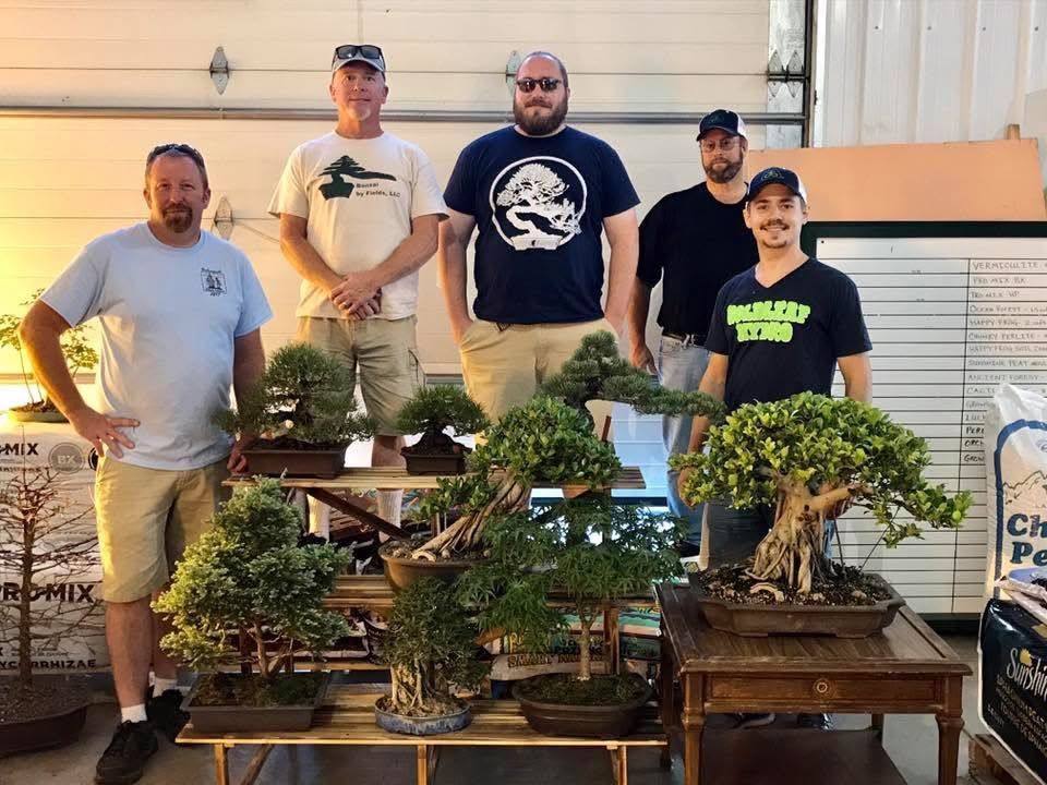 Five men stand behind rows of bonsai trees on display in a workshop or garage setting.