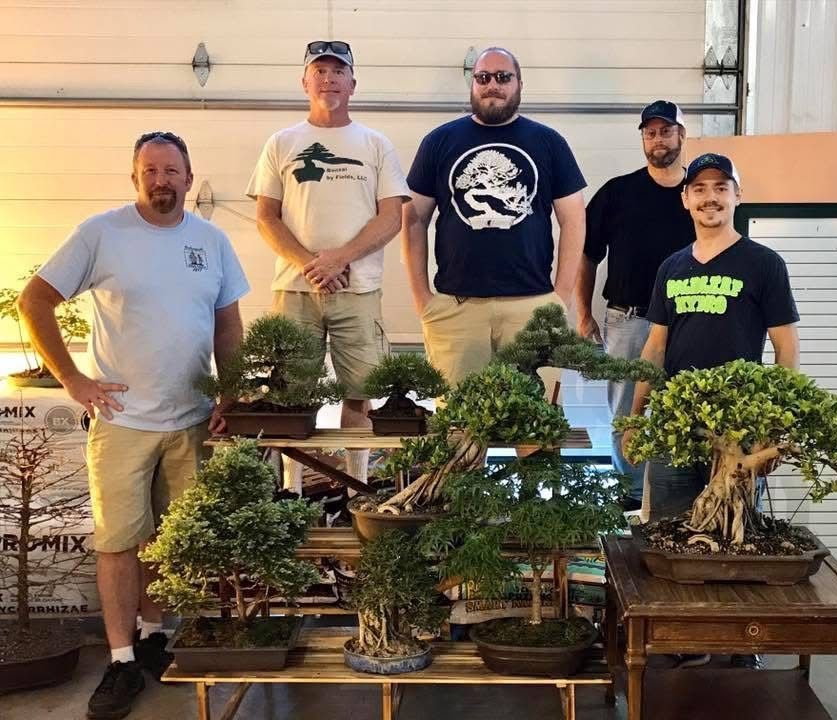 Five men pose behind several bonsai trees displayed on stands outdoors.