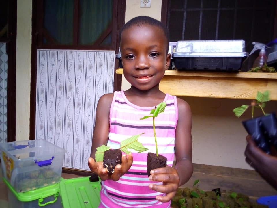 Child holding a potted seedling and a small plant start indoors, with gardening supplies nearby.