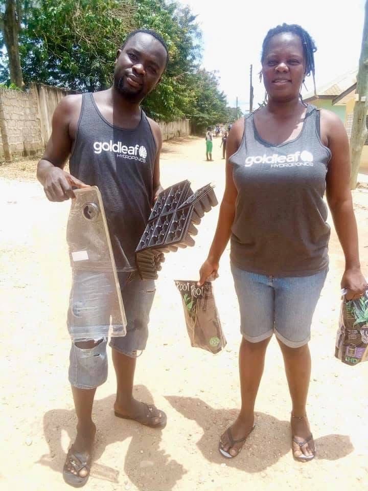 Two people in matching dark shirts stand outdoors on a sunny street, holding a metal grate and a small bag.