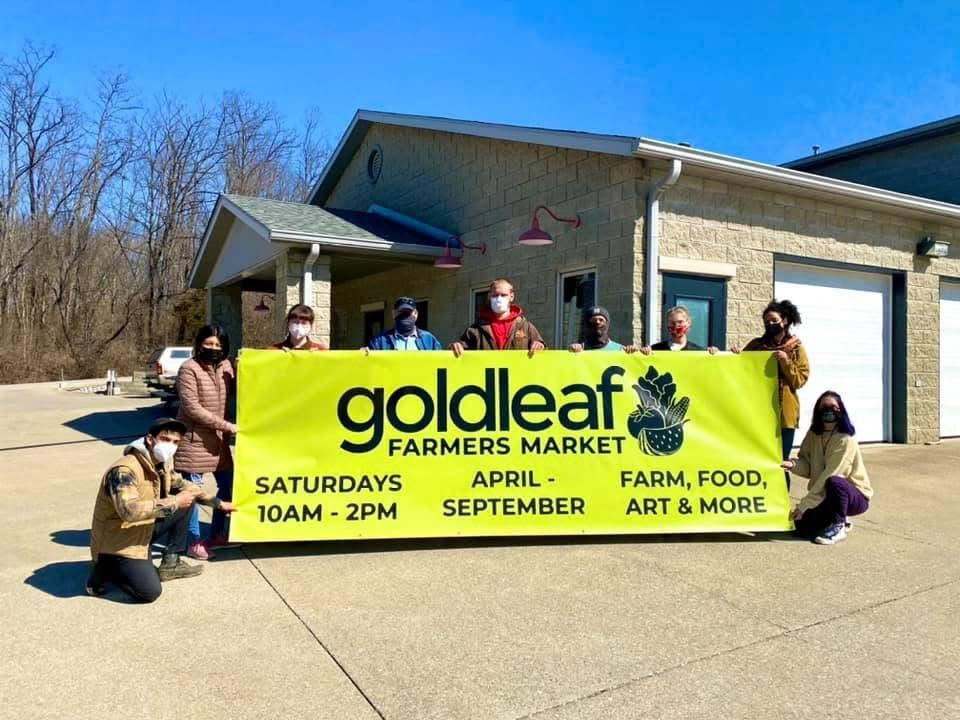 People holding a bright yellow goldleaf farmers market banner outside a building on a sunny day