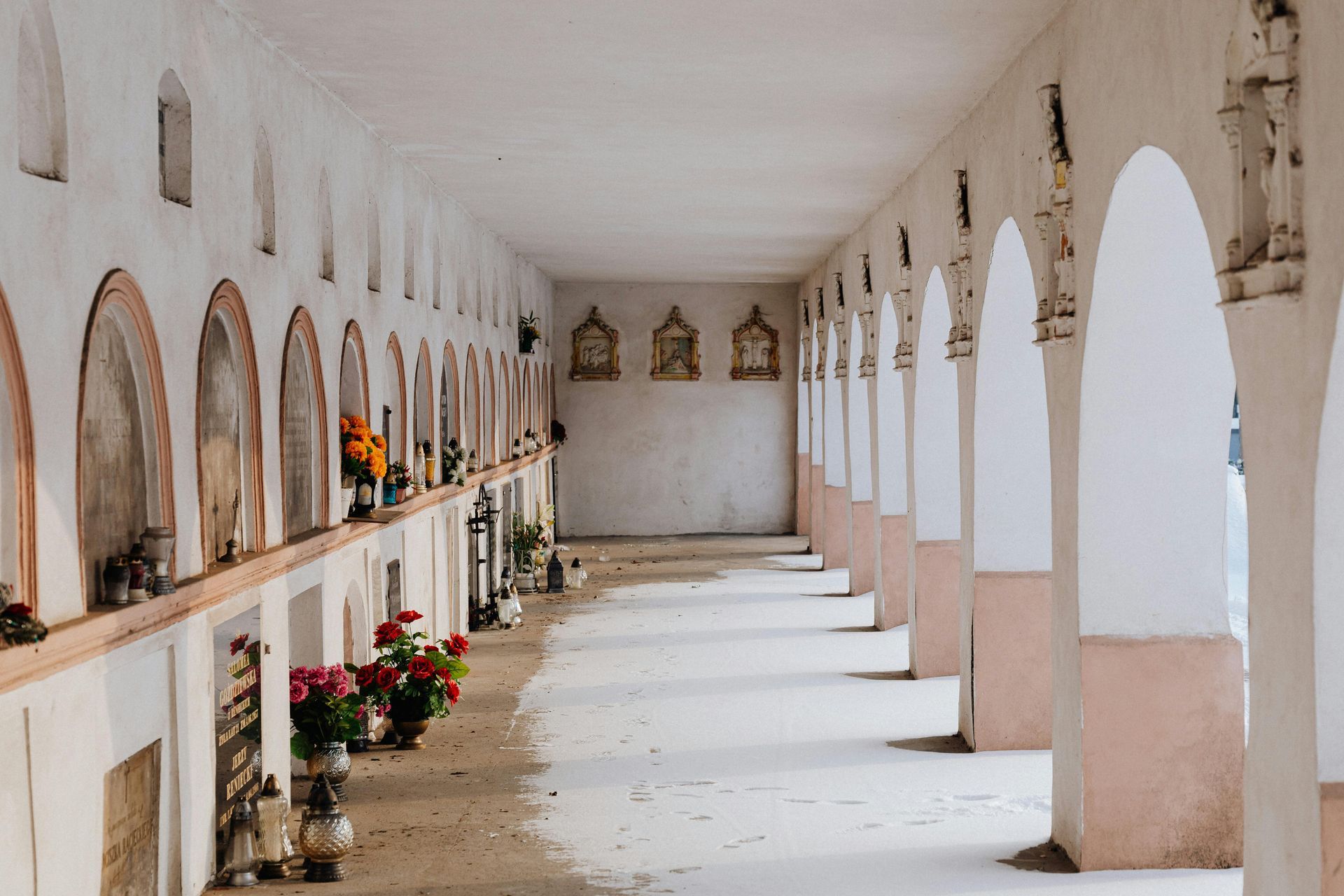 Lungo corridoio ad arco in un cimitero con nicchie che ospitano monumenti commemorativi; sono presenti fiori e candele.