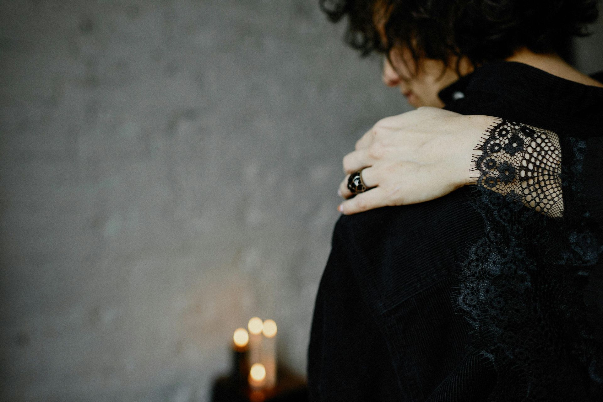 Una persona vestita di nero con maniche di pizzo si tocca la spalla vicino a candele accese.