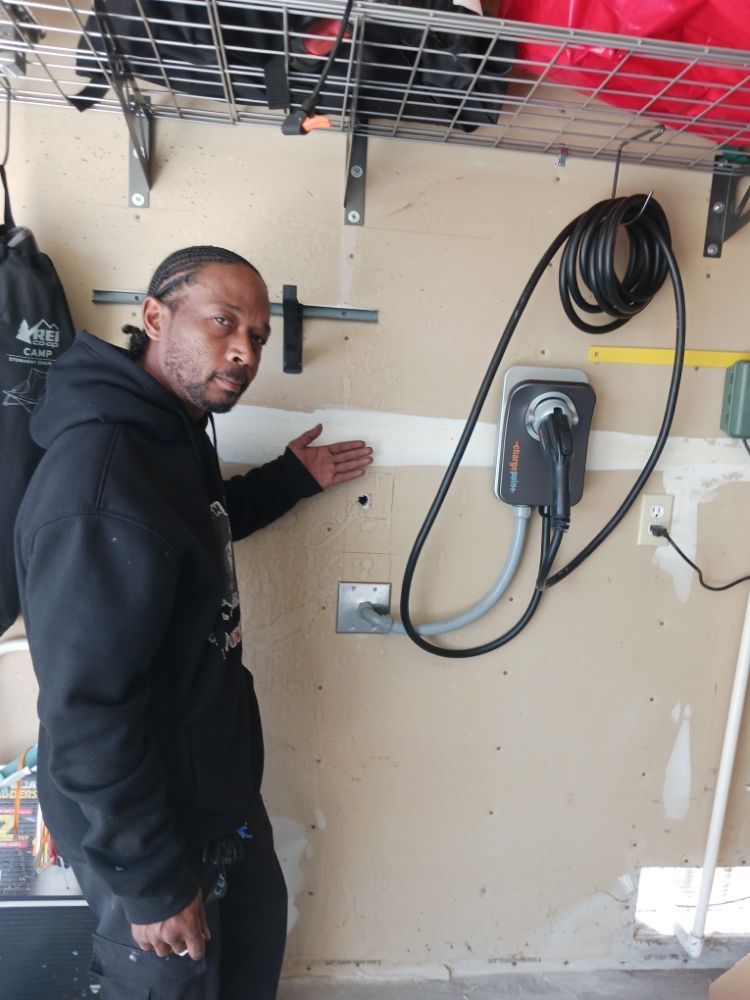 Man points to a wall with an EV charger in a garage. The wall is beige with some damage.
