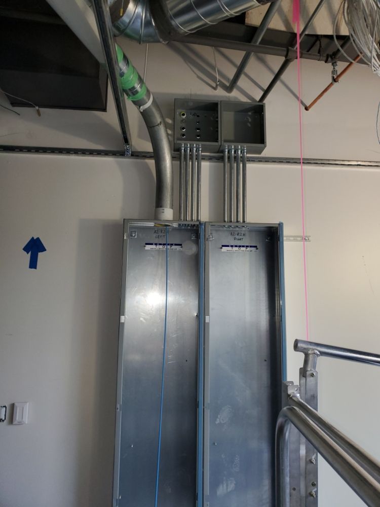 Metal electrical panel with conduits, attached to a white wall, near a ceiling with ductwork.