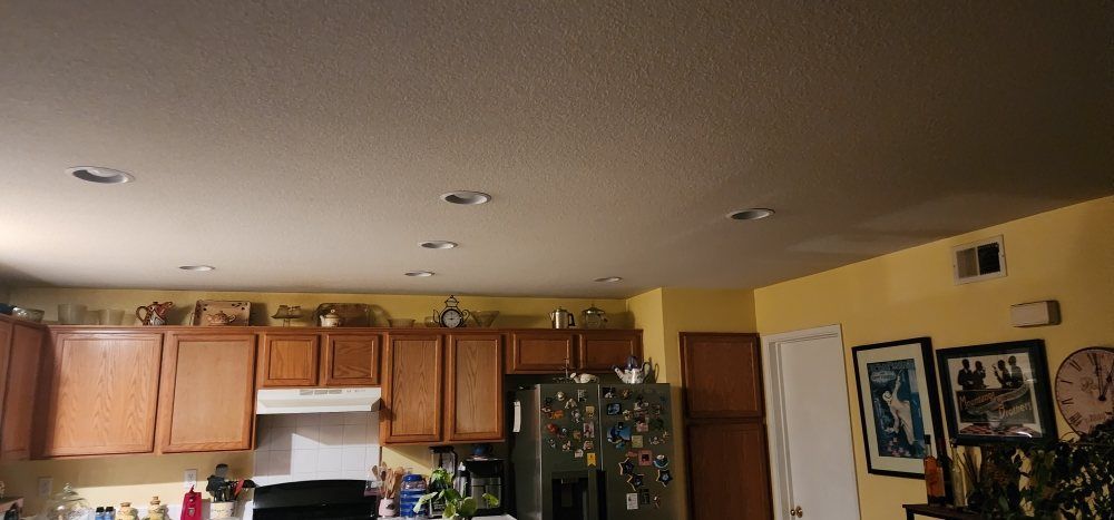 Kitchen interior with wood cabinets, yellow walls, and recessed ceiling lights. Refrigerator and artwork visible.