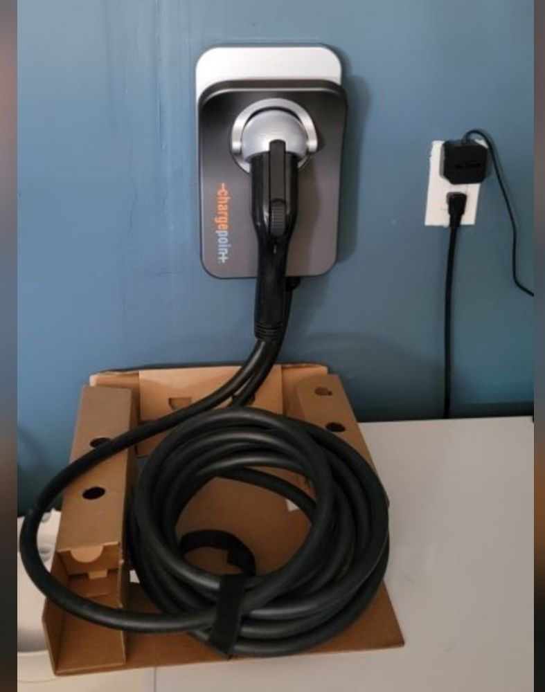 EV charger mounted on blue wall with cable coiled on a cardboard box. A power outlet is to the right.