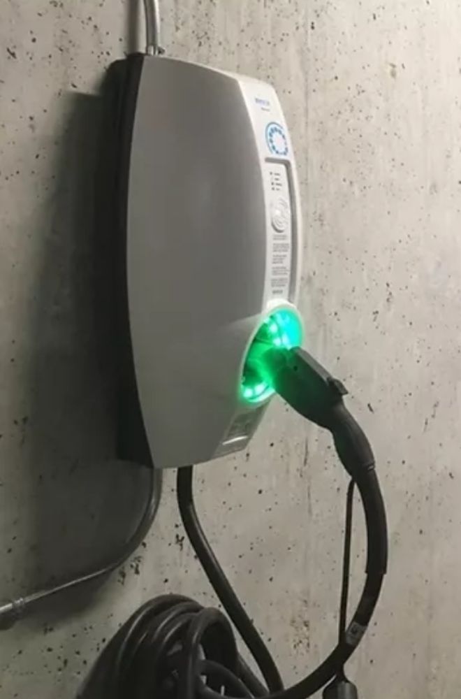 Electric vehicle charger mounted on a concrete wall, with green light indicating charging. Black cable coiled below.