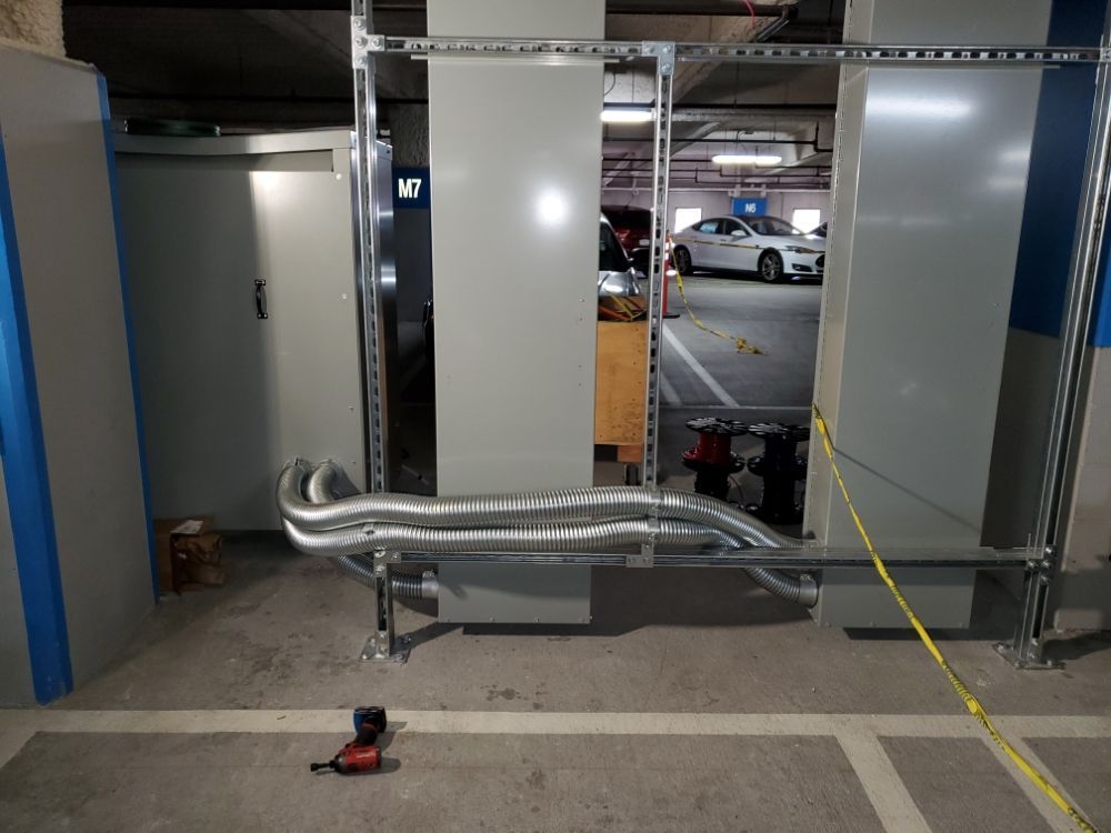 Metallic ductwork and gray panels in a parking garage. Silver pipes run near the floor, with a car visible in the background.