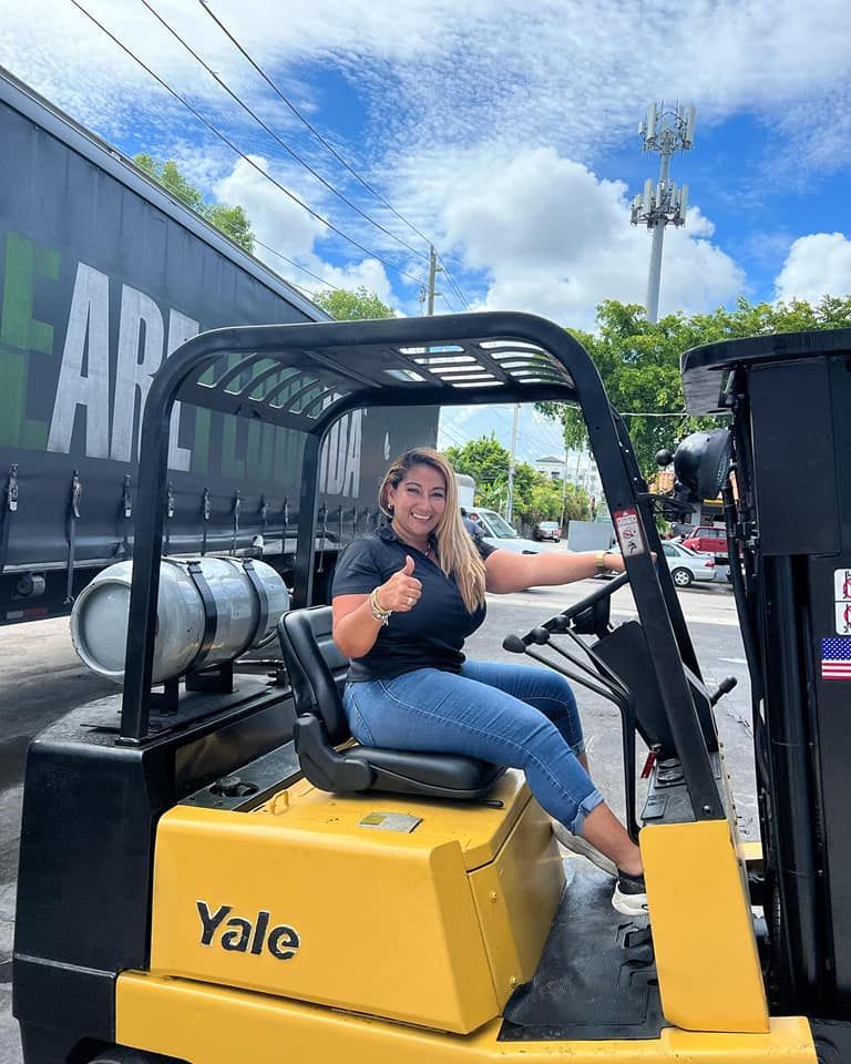 A woman is sitting on a yellow forklift and giving a thumbs up.