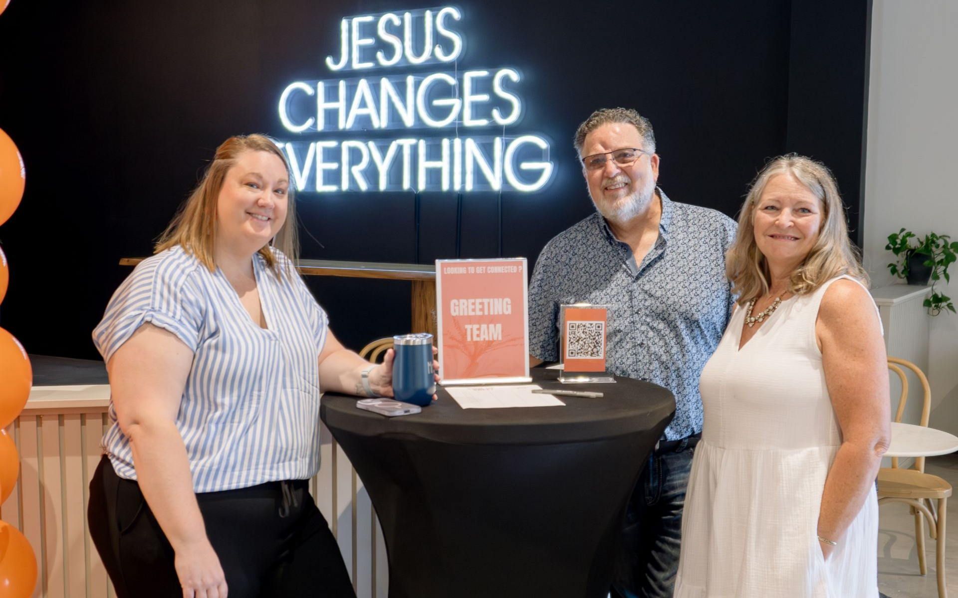 Three people smiling at a welcome table, under neon sign reading
