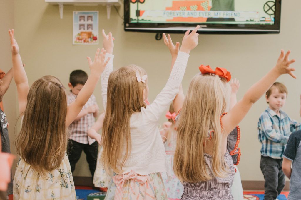 Children with arms raised, singing or dancing in a brightly lit room.