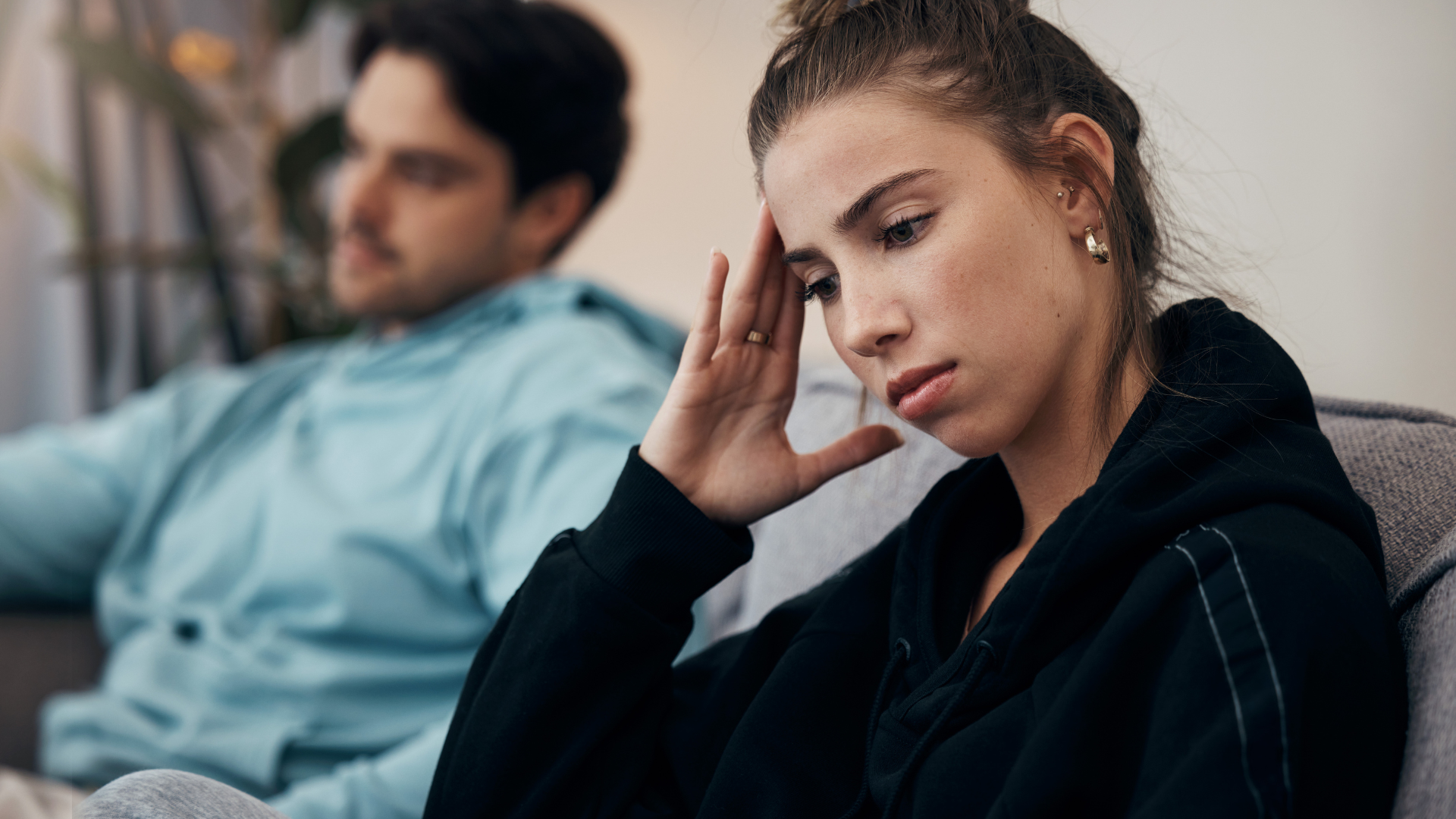 A man and a woman are sitting on a couch and the woman is holding her head.