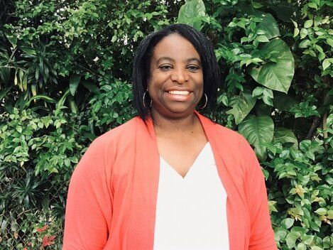 A woman in a red cardigan and white shirt is smiling in front of a bush.
