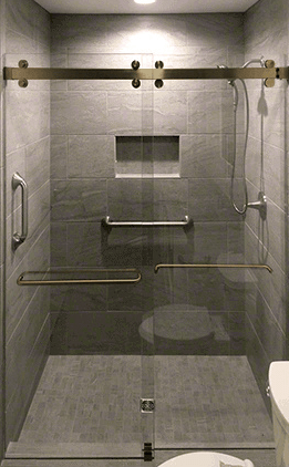Accessible shower with sliding glass door, gray tile walls, and brass grab bars.