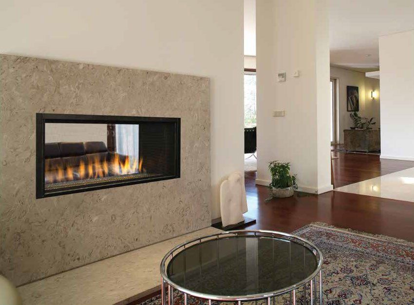 A modern horizontal gas fireplace built into a beige stone wall, viewed from a living room with a round coffee table.
