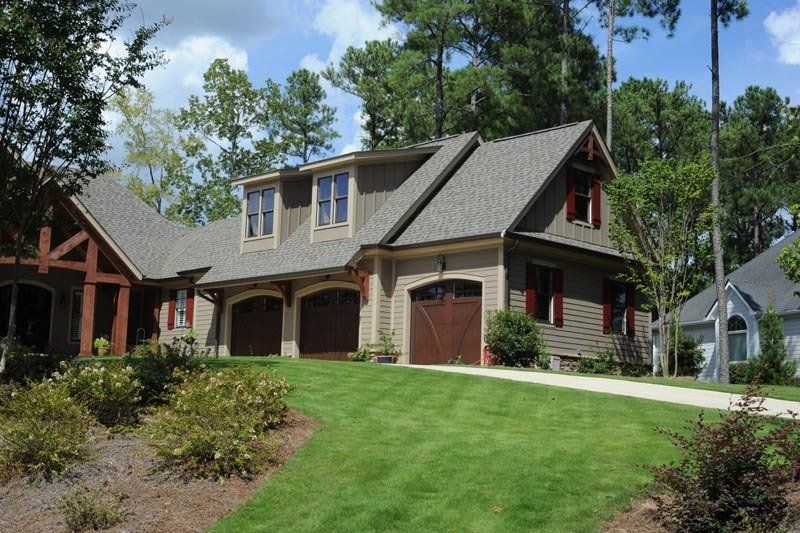A tan, multi-story house with a three-car garage and brown doors, surrounded by trees and a green lawn under a blue sky.