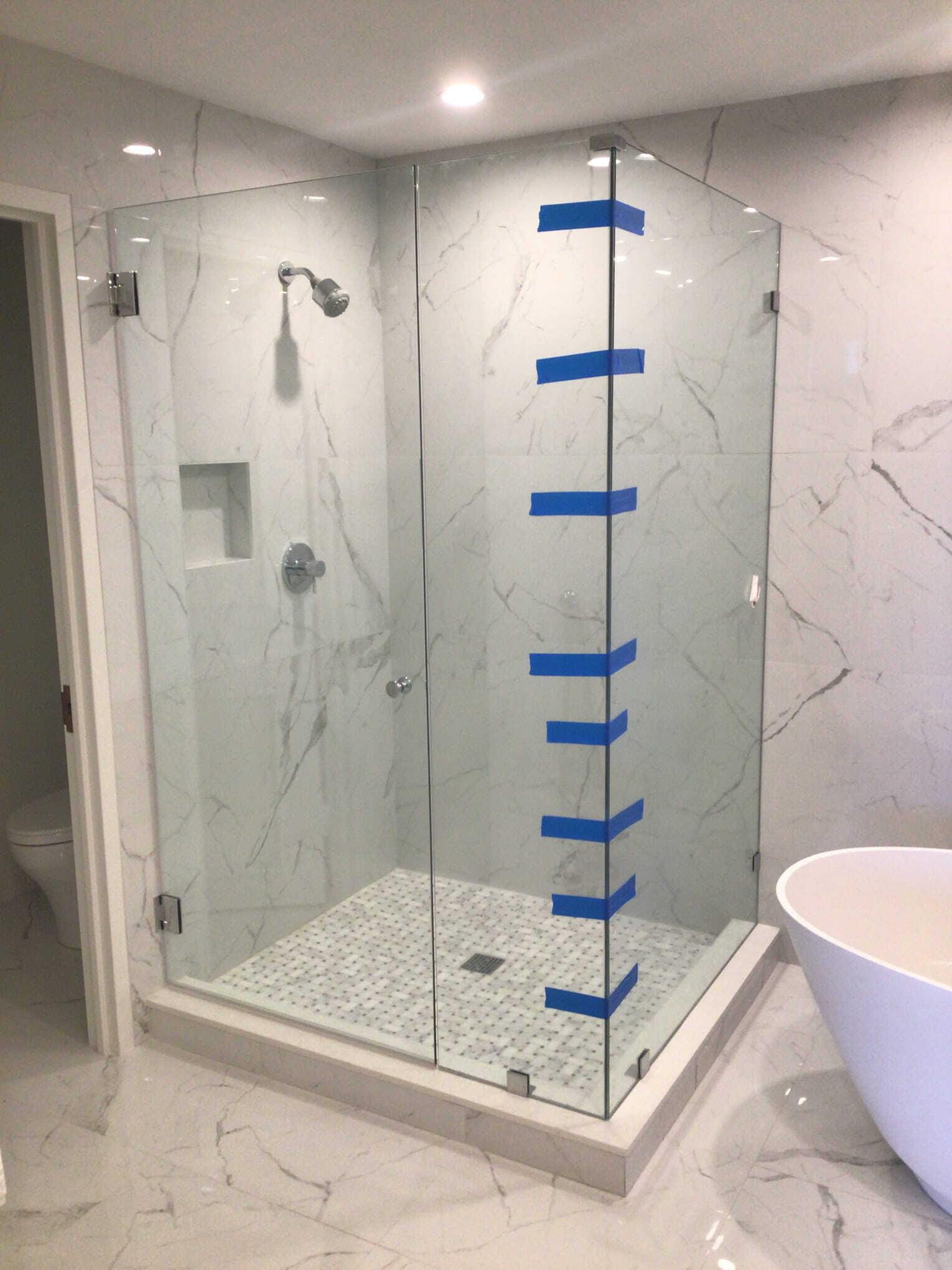 Glass shower enclosure with white marble tile, faucet, and showerhead. Blue tape on glass.