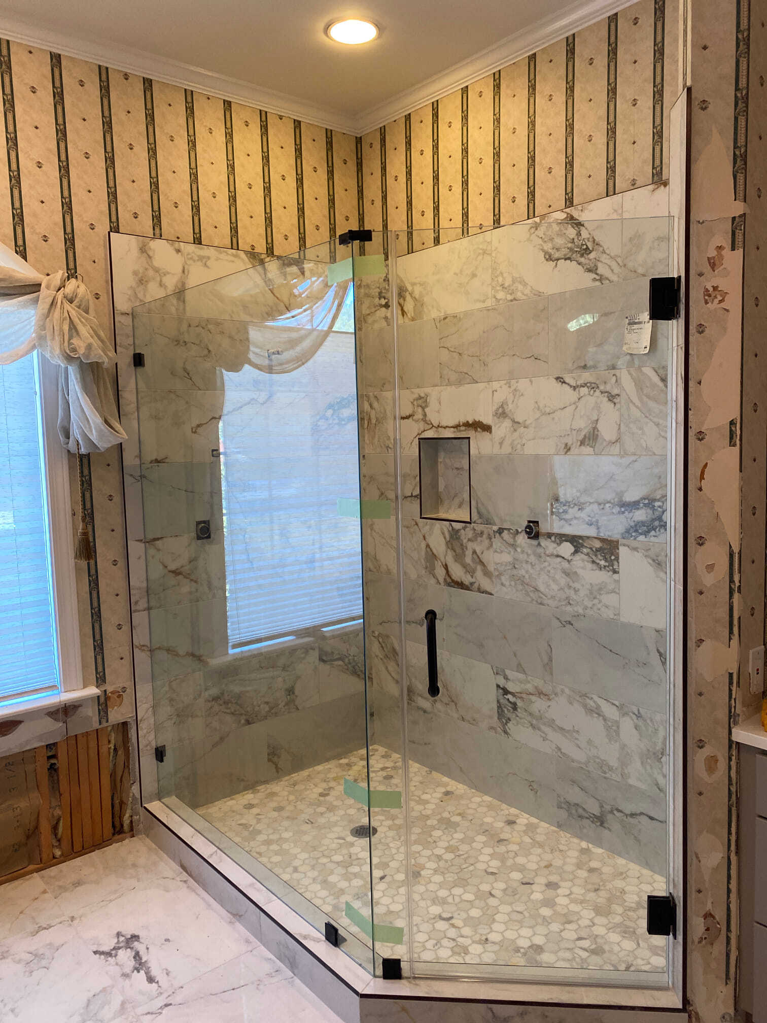 Glass shower with marble-look tile, black hardware, and pebbled floor. Window with blinds and patterned wallpaper visible.