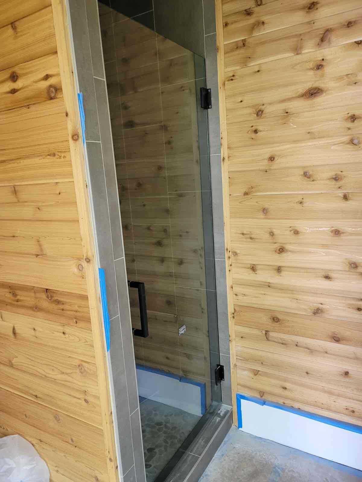Shower with glass door, cedar walls, gray tile frame. Blue tape at bottom.