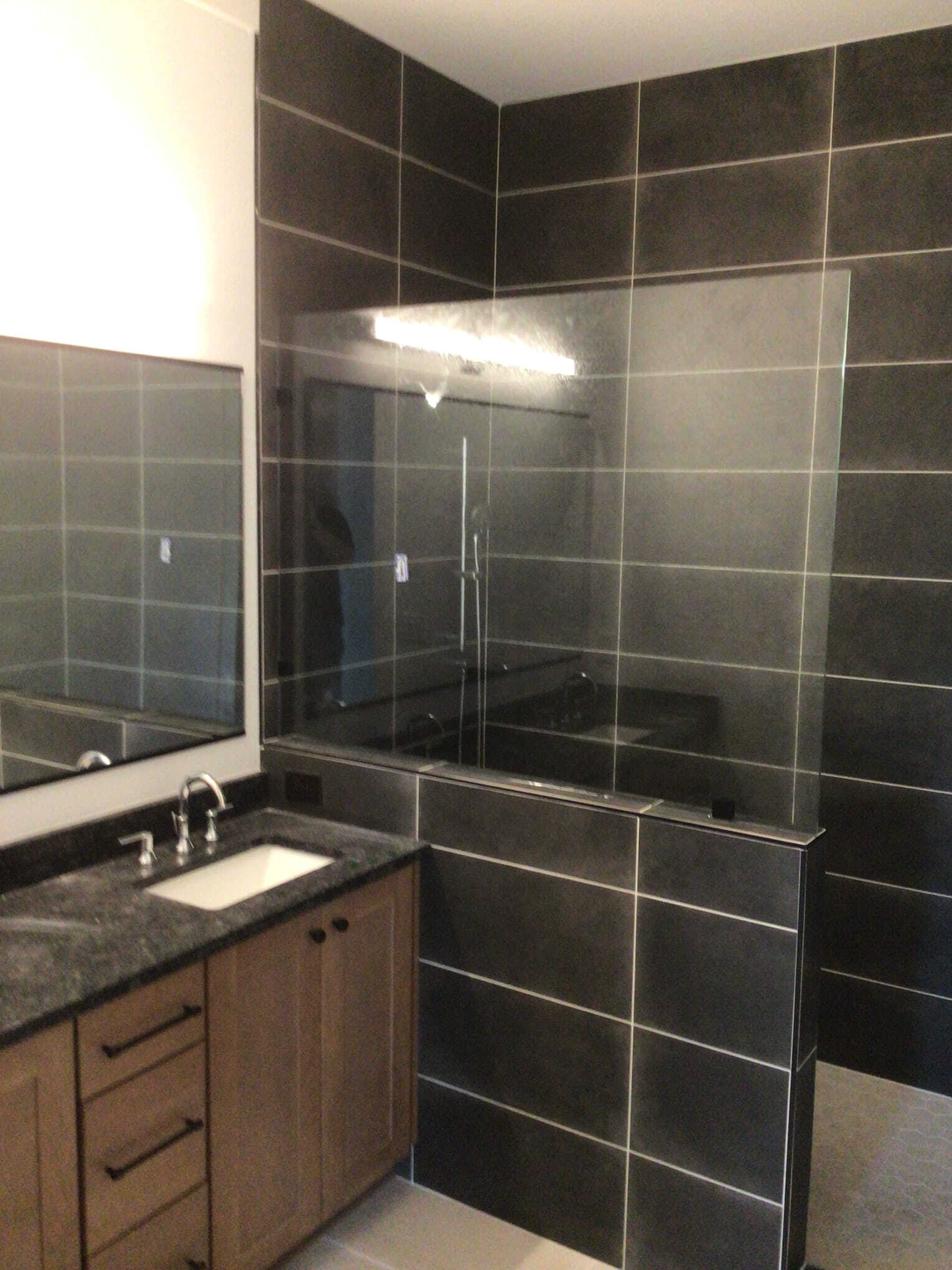 Bathroom with dark tiled walls, vanity, shower with glass panel, and light fixture.