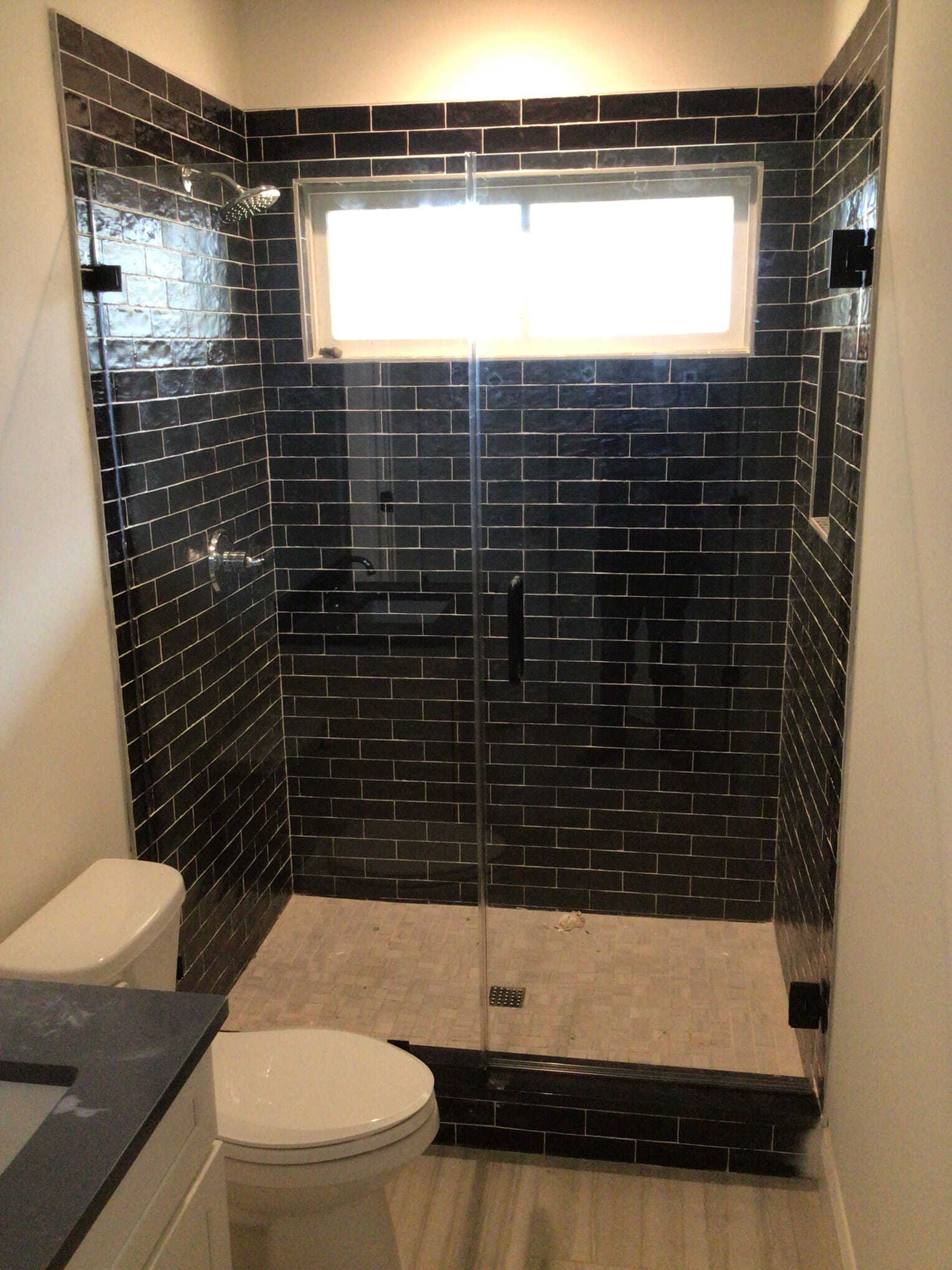 Black-tiled shower with glass doors and a window. A toilet is in the foreground.