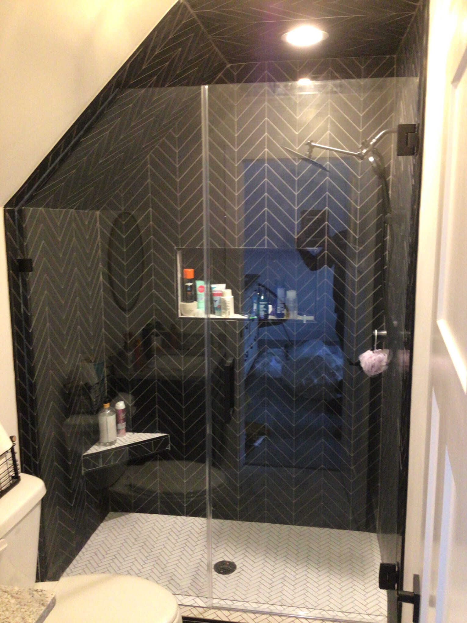 Modern shower with dark chevron-patterned tiles, glass door, white floor tiles, and a built-in shelf.