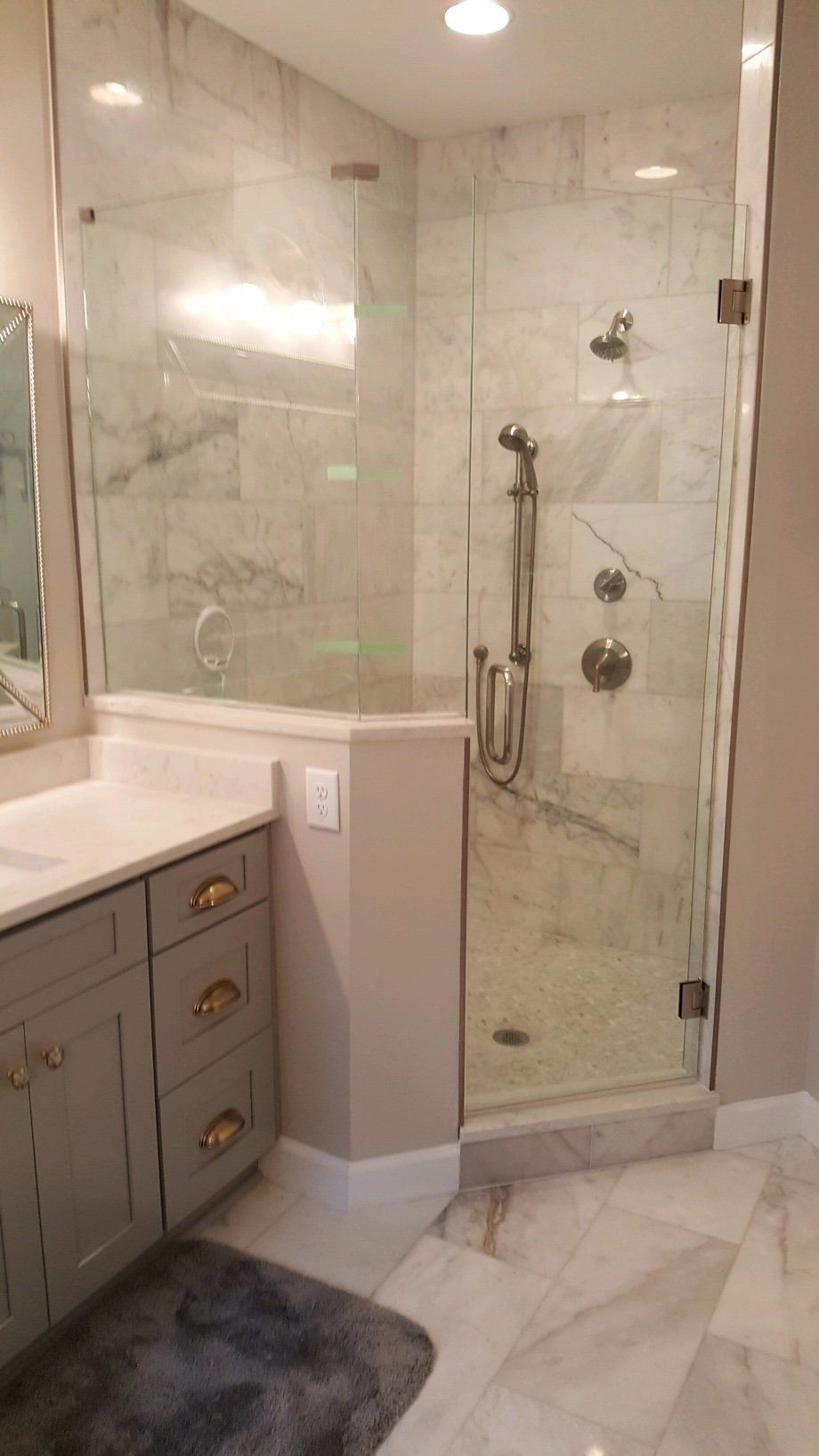 Corner glass shower with marble tile, gray cabinets, and a rug.