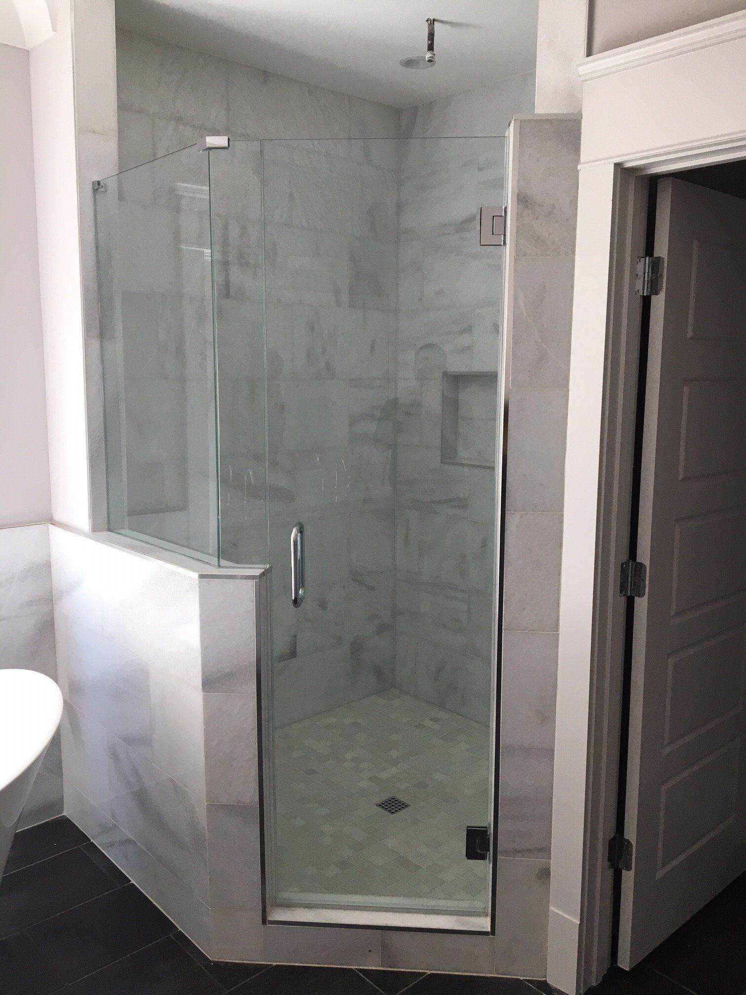 Glass-enclosed shower with white marble tile walls and dark floor. A door and an open doorway are visible.