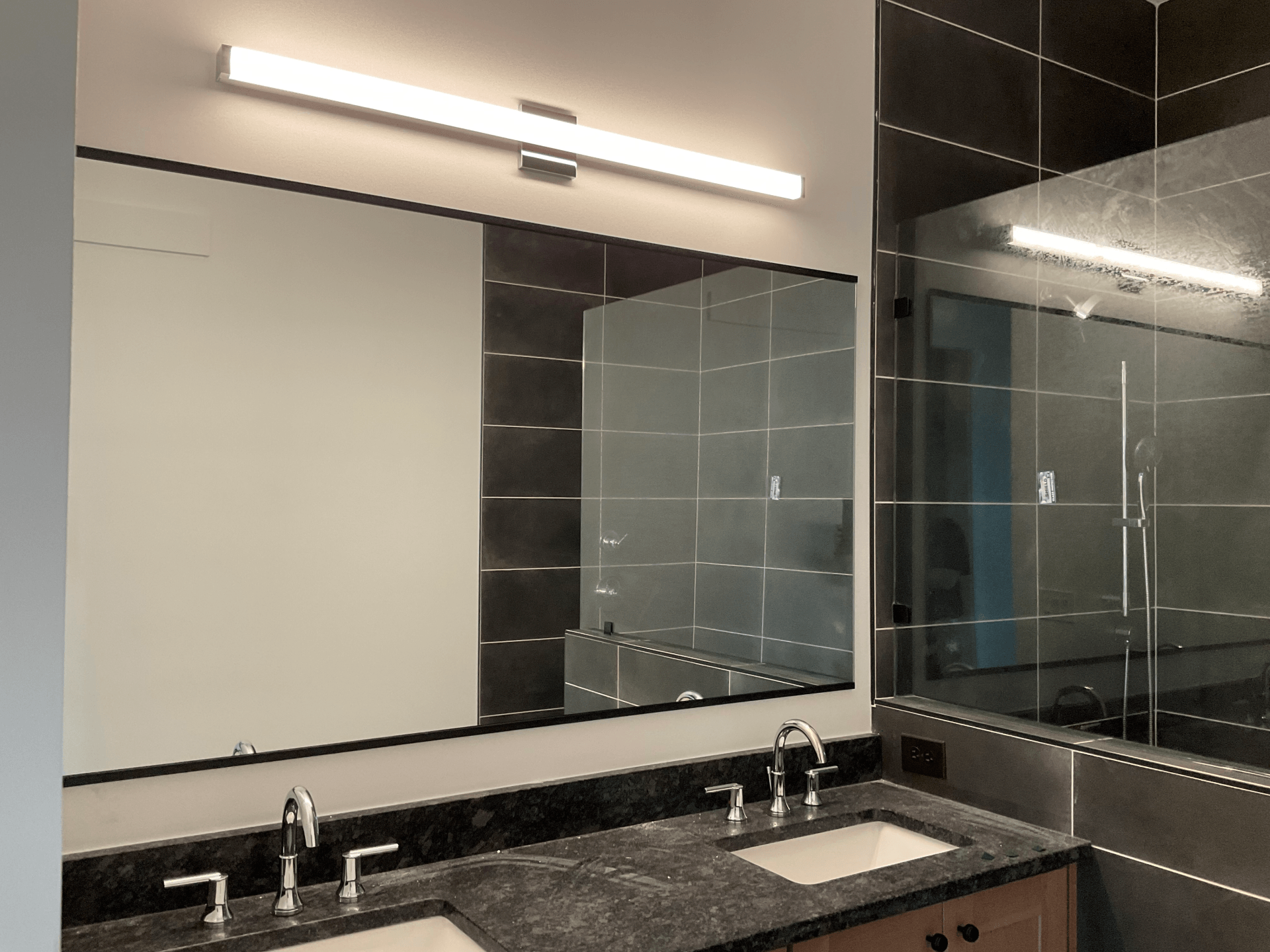Bathroom with large mirror, double sinks, dark granite countertop, and linear light fixture.