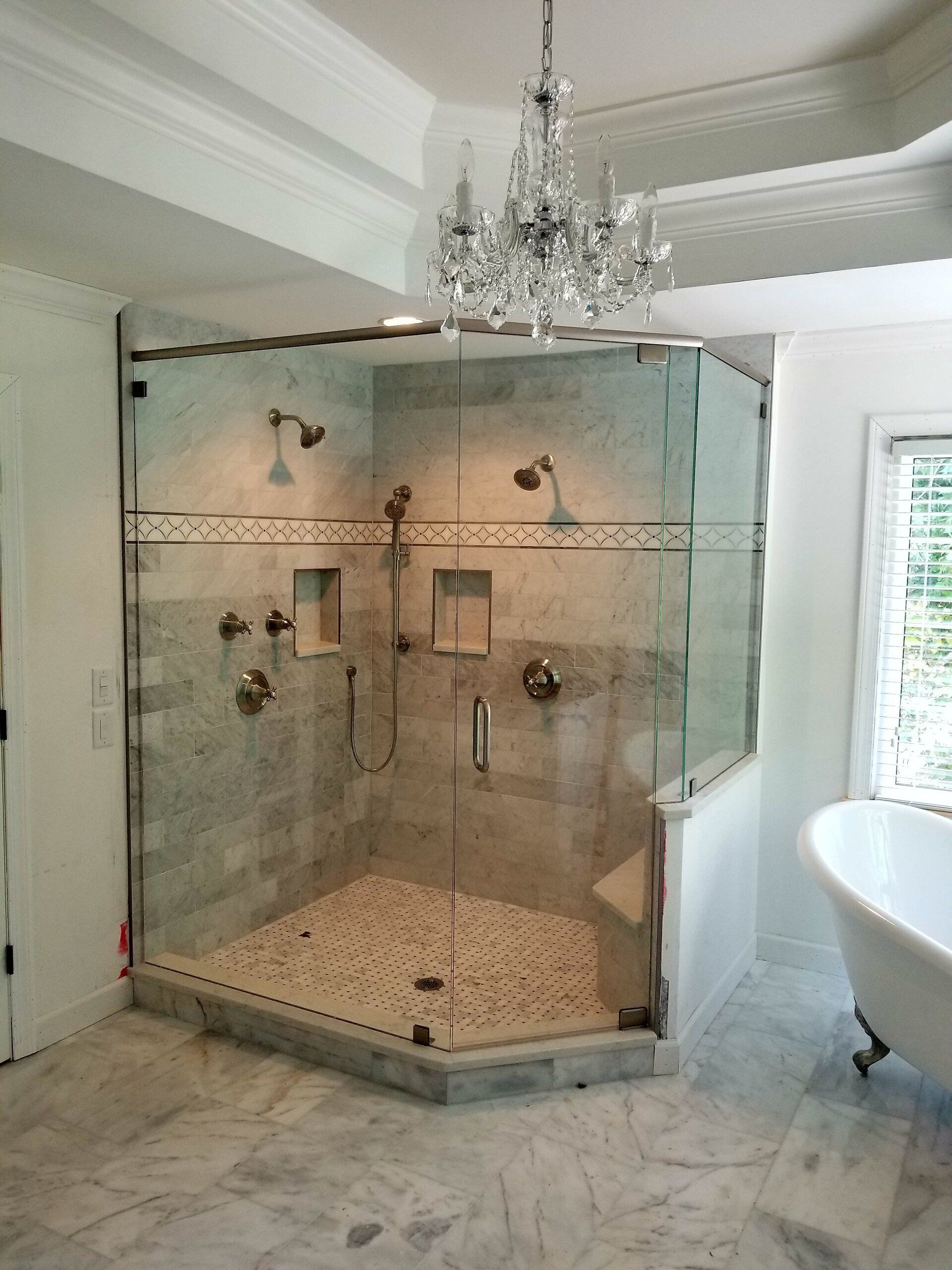 Luxurious marble-tiled shower with multiple showerheads and glass doors, lit by a crystal chandelier.
