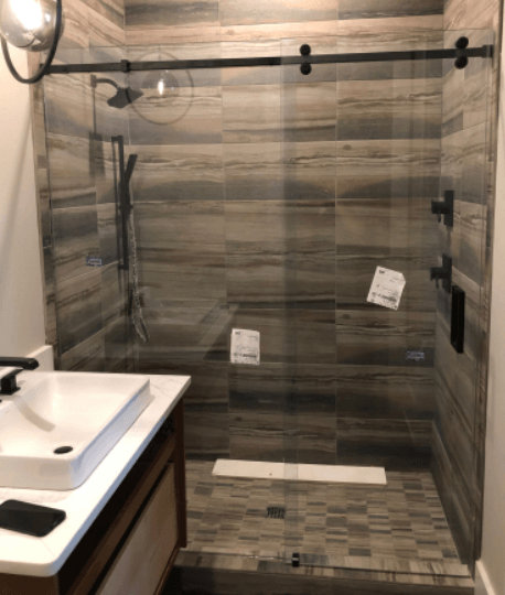 Shower stall with sliding glass door and dark fixtures, tiled with gray and brown horizontal stone-look tiles.
