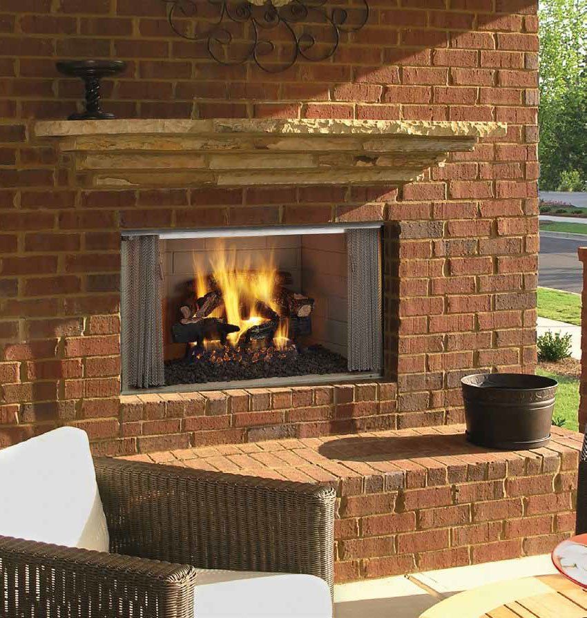 A brick outdoor fireplace with a burning gas fire, a stone mantel, and a wicker chair in the foreground.