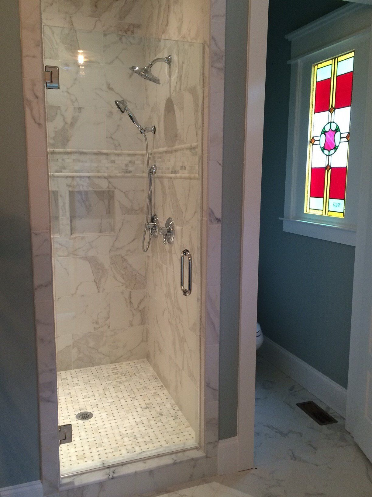 Marble shower with glass door. Mosaic floor tiles. Stained glass window in blue-walled bathroom.