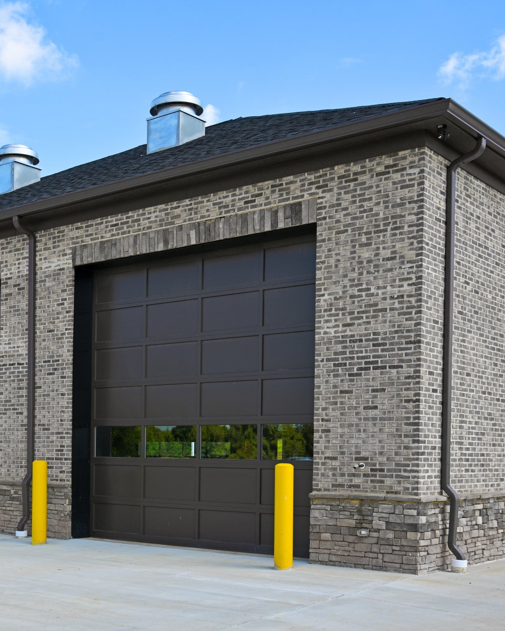A building exterior features a dark, multi-panel garage door, stone and brick siding, and two bright yellow bollards.