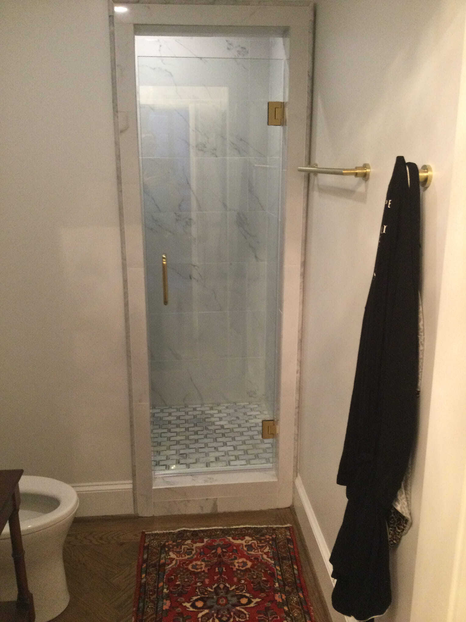 Bathroom with a glass shower door, gold fixtures, and a black robe hanging on a wall. A small rug is on the floor.