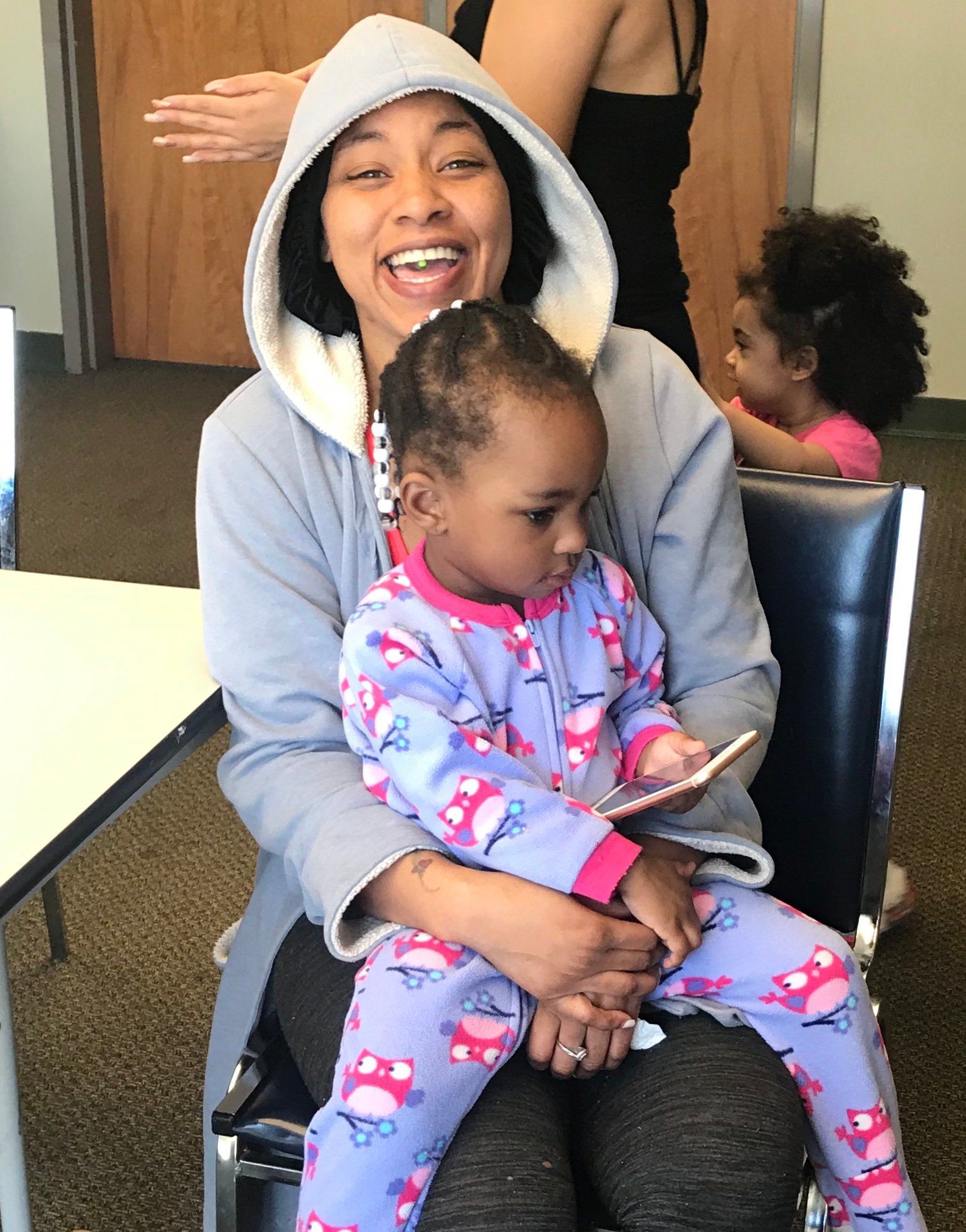 Smiling adult in gray hoodie holding a child in pink pajamas seated indoors on a chair