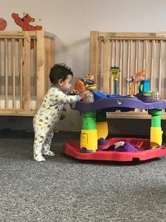 Toddler standing at a colorful activity table in a playroom with wooden cribs and gray carpet.