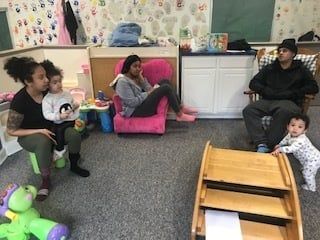 People sitting with babies and toddlers in a playroom, with toys and child-sized furniture.