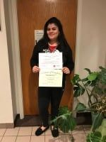 Woman standing indoors holding a certificate beside a potted plant.