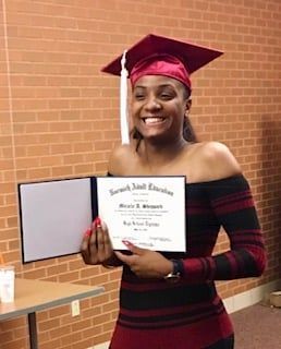 Graduate smiling and holding diploma, wearing maroon cap and gown in front of a brick wall.